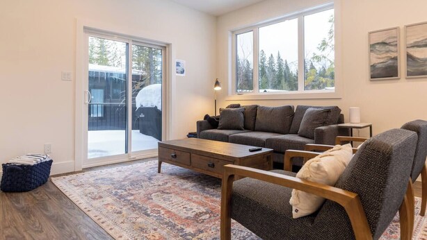 A spacious living room with a large sofa, a coffee table, and a rug. There are large windows providing natural light and a view of trees outside. - Gorgeous 2 Bedroom with Private Hot Tub. A/C (Fernie)
