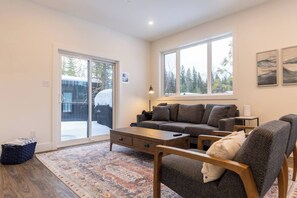 A spacious living room with a large sofa, a coffee table, and a rug. There are large windows providing natural light and a view of trees outside.