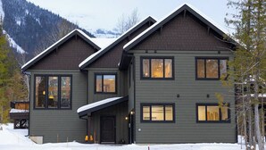 A two-story house with a dark exterior, large windows, and a snow-covered roof. - Gorgeous 2 Bedroom with Private Hot Tub. A/C (Fernie)