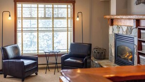 Ein gemütliches Wohnzimmer mit Kamin, zwei Ledersesseln, einem kleinen Tisch und einem großen Fenster mit Jalousien.