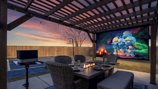 Eine Terrasse im Freien mit Esstisch, Stühlen, einem Fernseher auf einem Stativ und einer Feuerstelle.