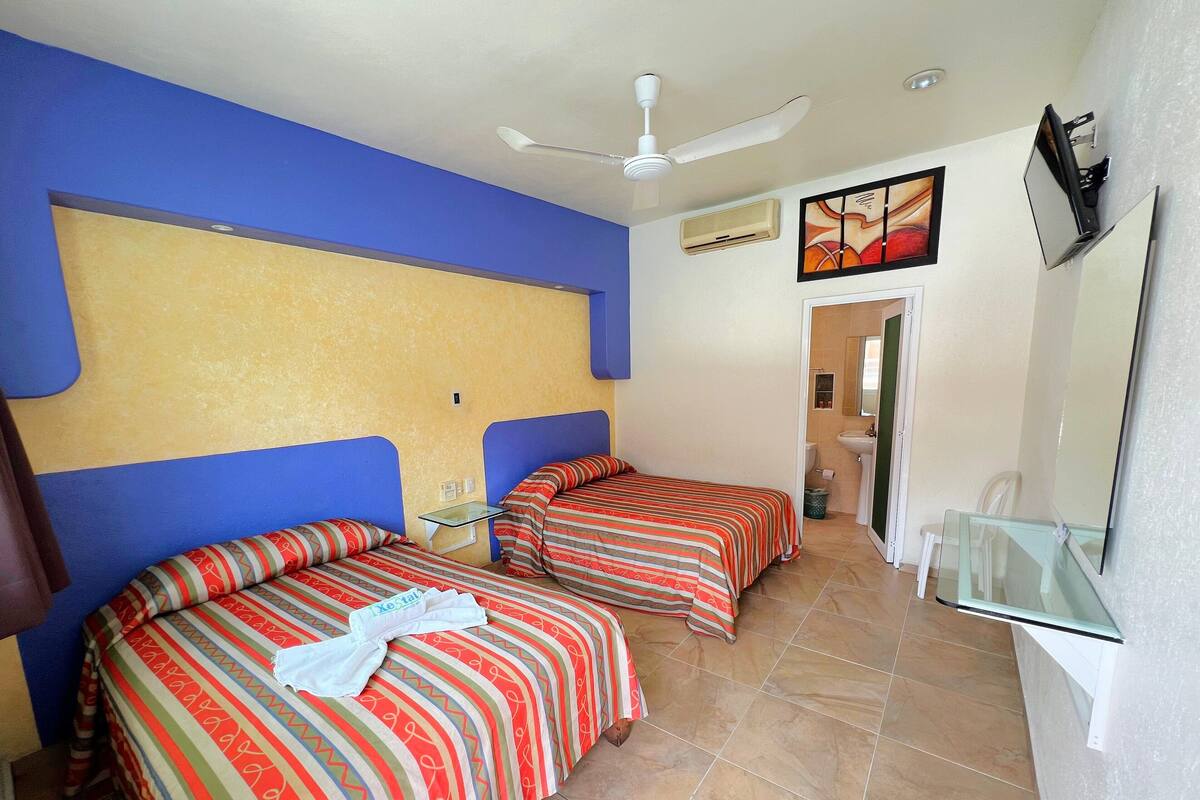 Une chambre d’hôtel avec deux lits, un ventilateur de plafond, un appareil de climatisation, une salle de bain avec un lavabo et des toilettes, et un téléviseur fixé au mur.
