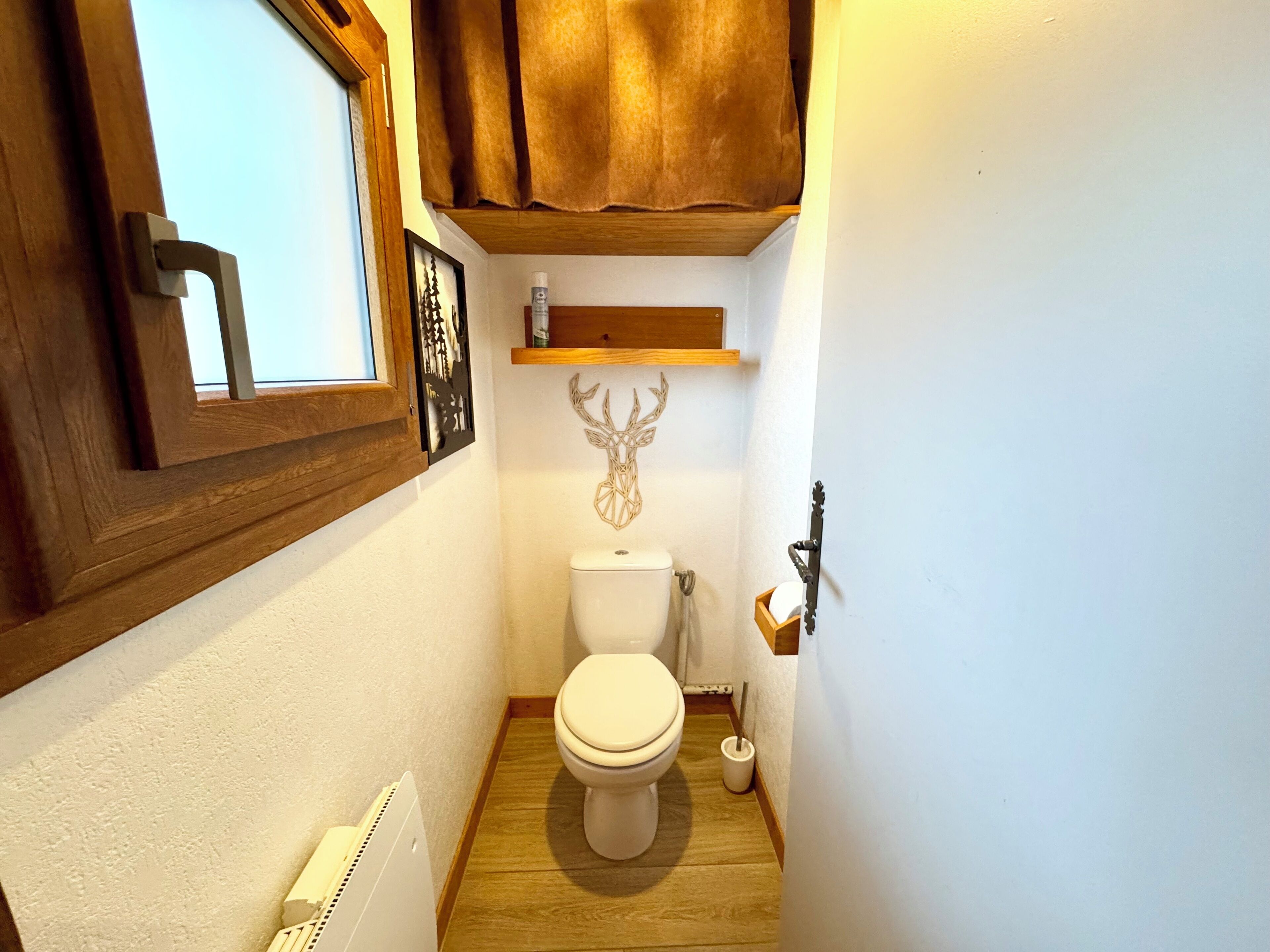 Un baño pequeño con suelo de madera, un inodoro blanco, un cuadro en la pared y un estante con una decoración de cabeza de venado.