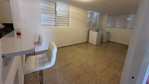 Quarto com cozinha compacta, um banquinho de bar, uma cadeira, máquina de lavar e luminária no teto.
