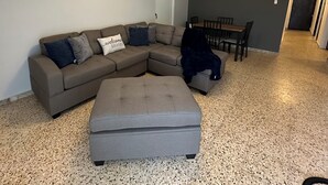 Uma sala de estar com sofá modular, um banquinho cinza, uma mesa de jantar com cadeiras e uma área de cozinha ao fundo.