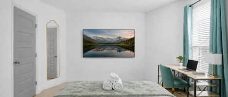 Un dormitorio con una cama, un escritorio con una computadora portátil, una silla, un cuadro de un paisaje montañoso y una puerta.