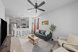 A modern living room with a sofa, coffee table, and a TV. There is a kitchen area with a refrigerator, microwave, and bar stools. A ceiling fan is visible, and the room has a neutral color scheme. - Ideal for long-term stays-available for 14 days and Beyond. (Temple)