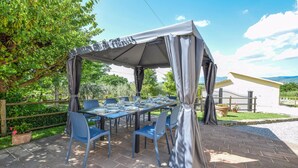 An outdoor dining area with a covered table and chairs, surrounded by greenery and a view of a building in the background.