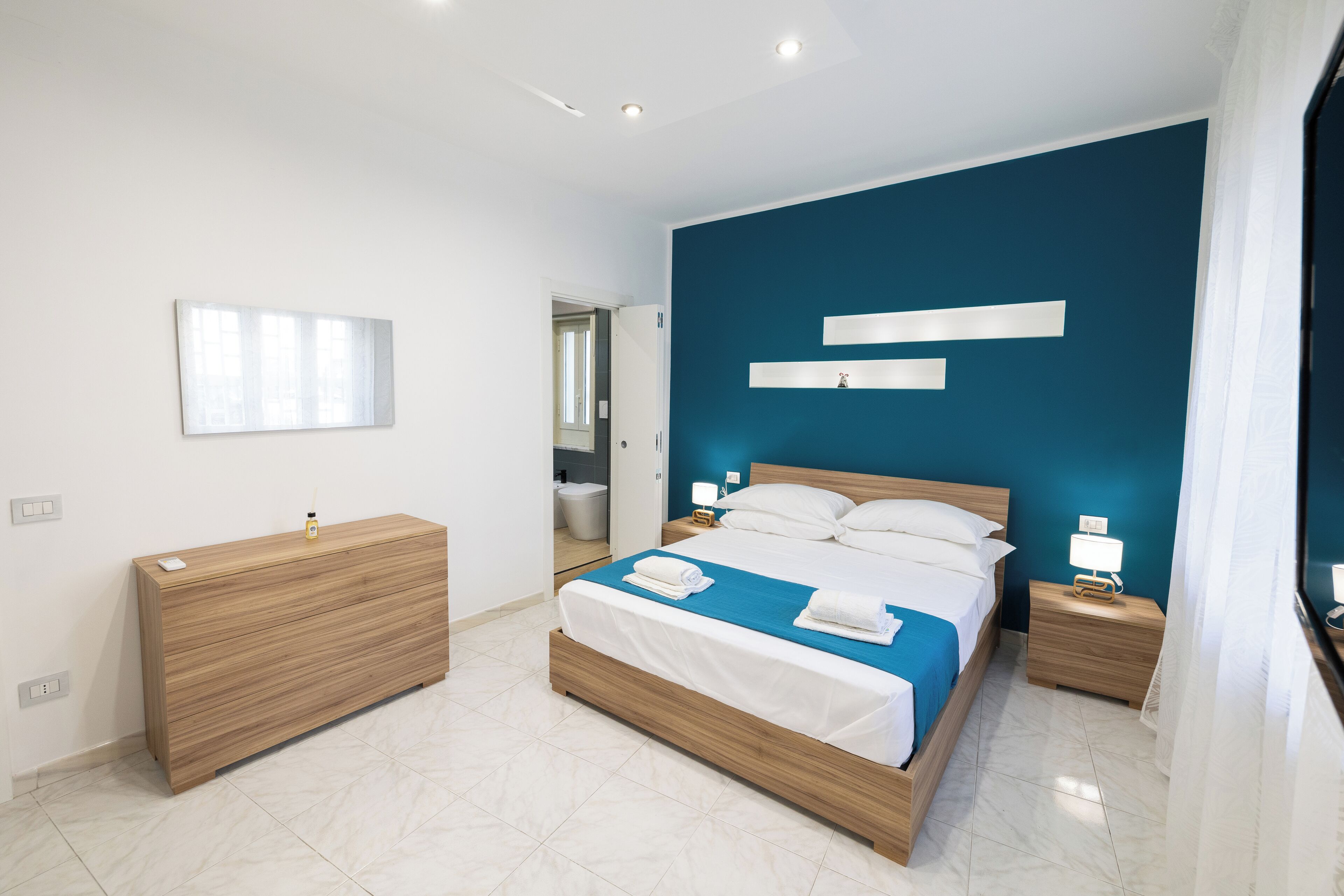A modern bedroom with a large bed, wooden dresser, and a blue accent wall.
