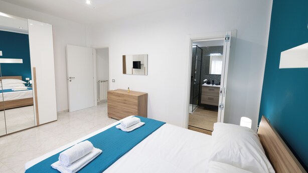 Une chambre d’hôtel moderne avec un grand lit, une commode en bois, un miroir et une salle de bain visible à travers une porte ouverte.