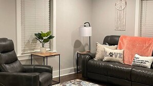 Ein Wohnzimmer mit einem schwarzen Ledersofa, einem schwarzen Lederfauteuil, einer Stehlampe, einer Wanddeko aus Makramee und einem Deckenventilator.