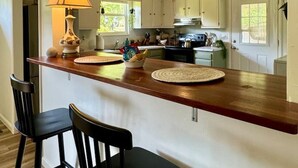 Una cocina con armarios blancos, encimera de madera, dos taburetes negros y una lámpara.