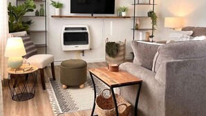 Una sala moderna con un sofá gris, una mesa de centro de madera, un televisor montado en la pared, un ventilador de techo y una mesita auxiliar con una lámpara.