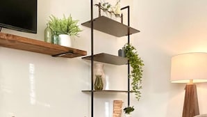 Una sala de estar con piso de madera, un sillón gris, una estantería de metal negro con plantas, una cesta tejida y una lámpara sobre una mesita.
