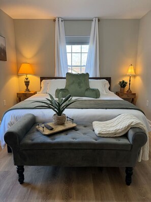 Une chambre à coucher avec un lit, deux tables de chevet avec des lampes, un fauteuil vert, une petite table avec une plante et un banc gris.