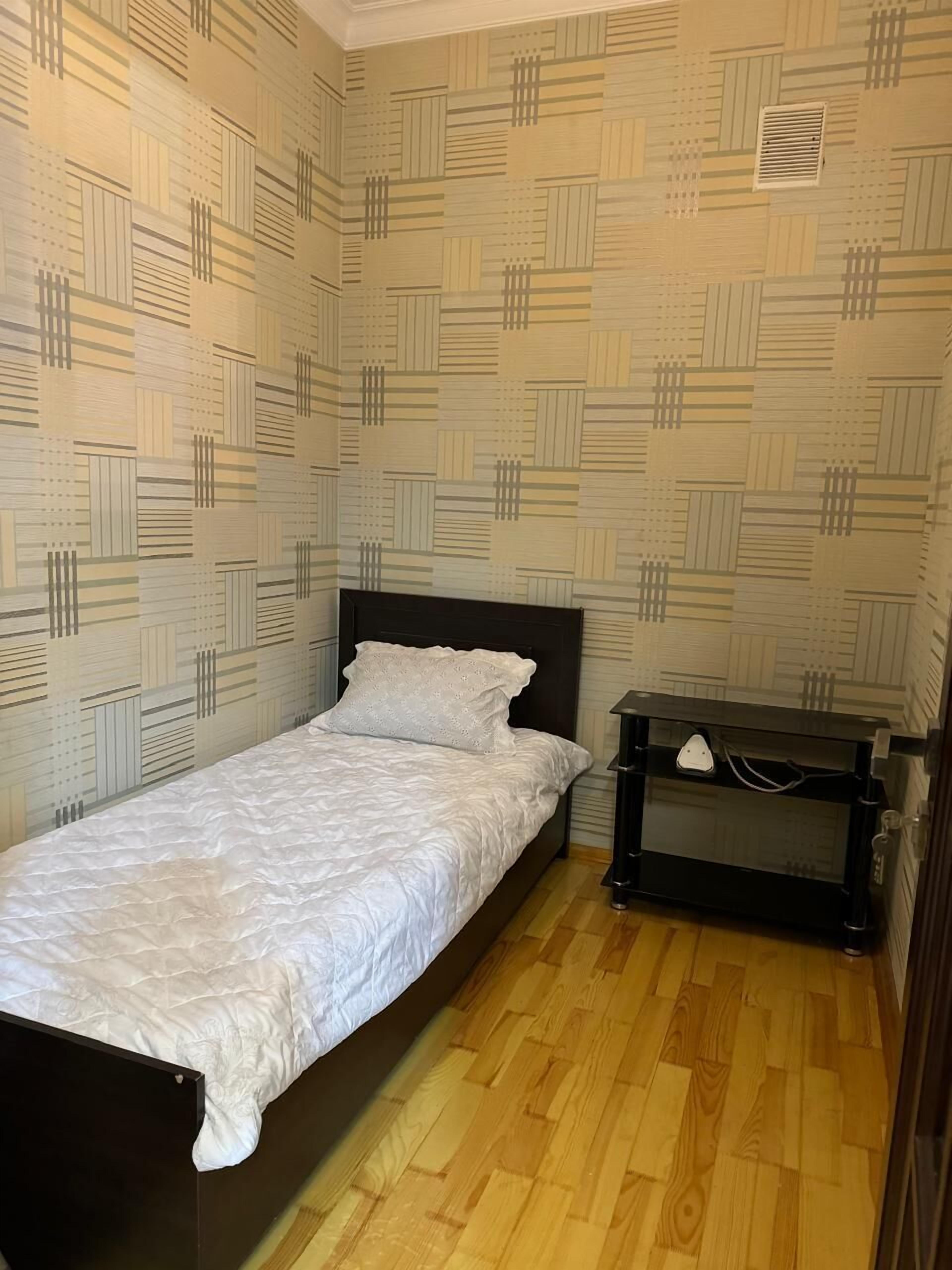 A small, single-bed room with a patterned wallpaper, a wooden floor, and a bedside table with a lamp.