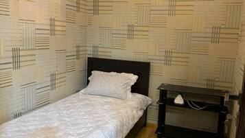A small, single-bed room with a patterned wallpaper, a wooden floor, and a bedside table with a lamp.