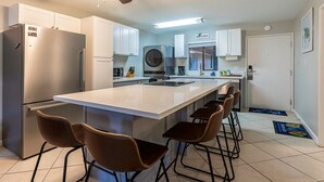 A modern kitchen with a central island, bar stools, a ceiling fan, and a sign reading "BEACH HOUSE".
