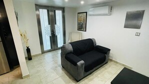 Una sala de estar con un sofá gris, una silla blanca, un cuadro en la pared y un equipo de aire acondicionado.