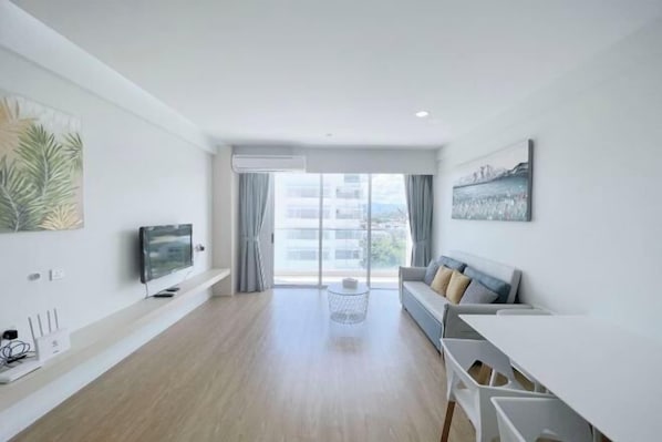 A modern living room with a flat-screen TV, a sofa, a dining table, and a large window with curtains.