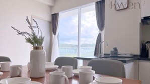A dining area with a table set for four, white tableware, a vase with greenery, and a view of the sea through large windows.