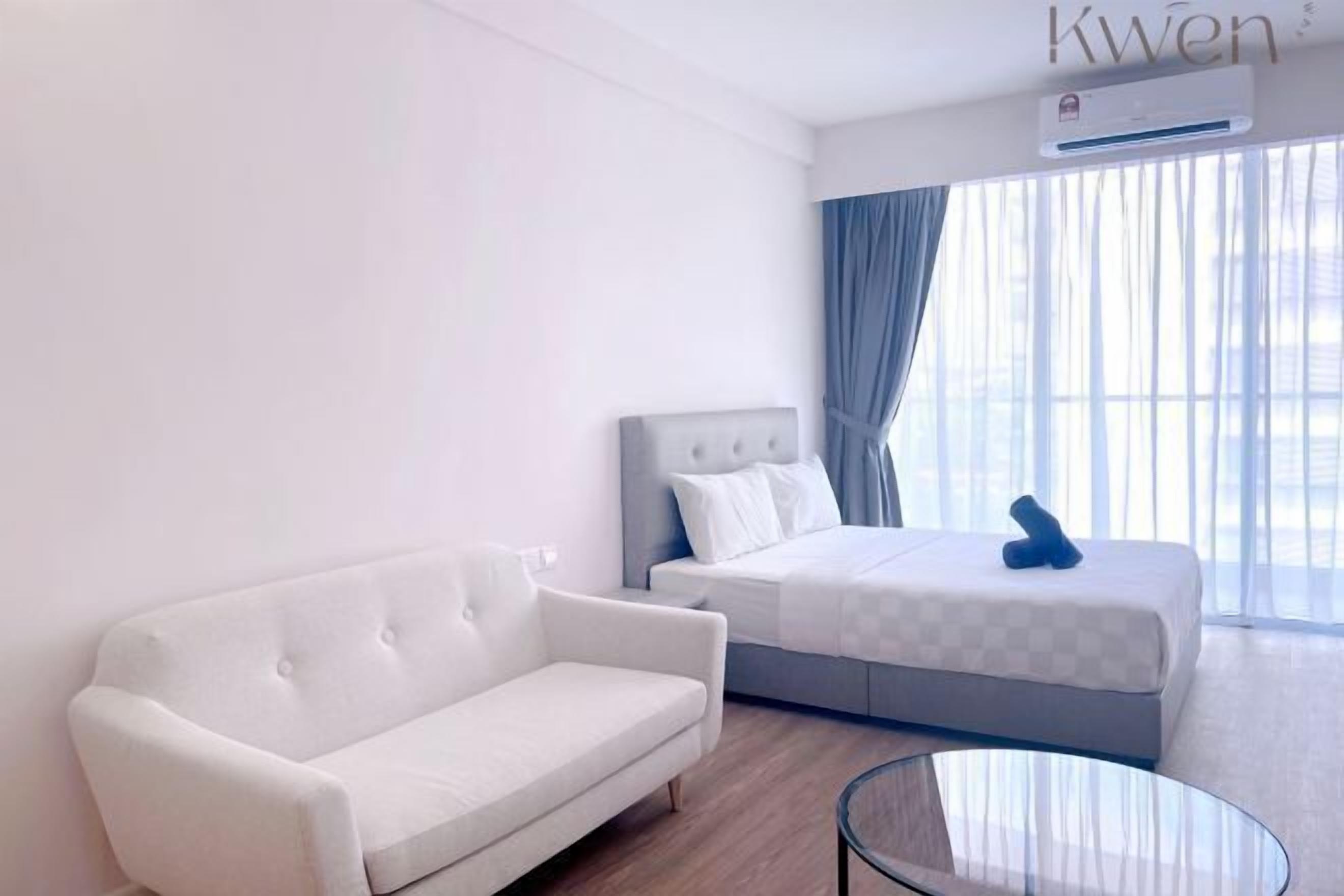 Een hotelkamer met een witte bank, een bed met een geruit dekbedovertrek, een glazen salontafel en een raam met gordijnen.
