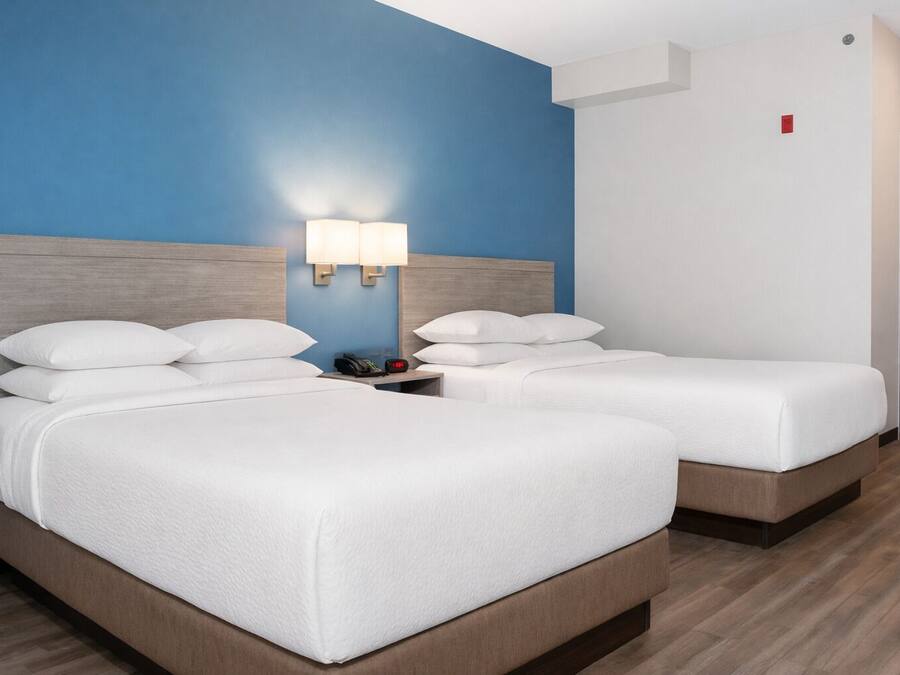 A hotel room with two beds, a nightstand with a lamp, and a door with a red emergency exit sign.
