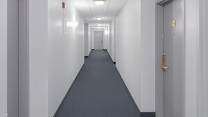 Un long couloir étroit aux murs blancs et au tapis sombre. Des portes se trouvent de part et d’autre, et un détecteur d’incendie est visible sur le mur gauche.