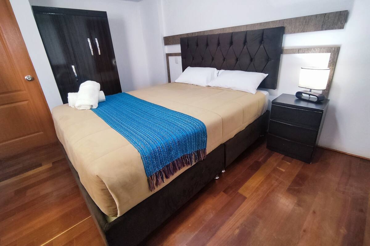 Un lit bien fait, avec une couverture bleue, une tête de lit en bois et une table de chevet surmontée d’une lampe.