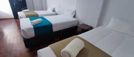 Habitación de hotel con tres camas, cada una con ropa blanca y una toalla enrollada. El suelo es de madera y al fondo se ve un armario oscuro.