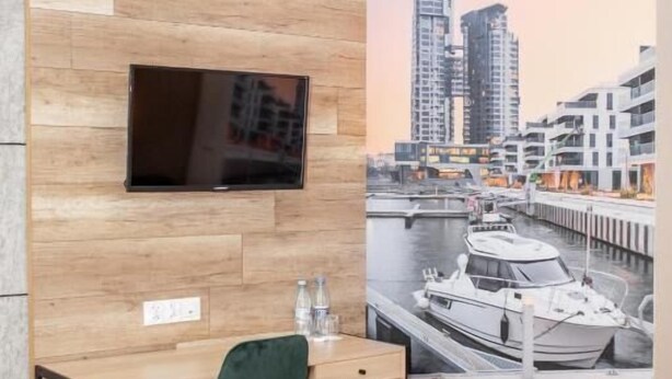 Une chambre d’hôtel avec un mur en bois, un téléviseur à écran plat fixé au-dessus d’un bureau, une chaise verte et un lit avec du linge de lit blanc.