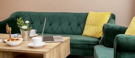 A green sofa with yellow cushions, a wooden coffee table with a laptop, a bowl of food, and a white cup.