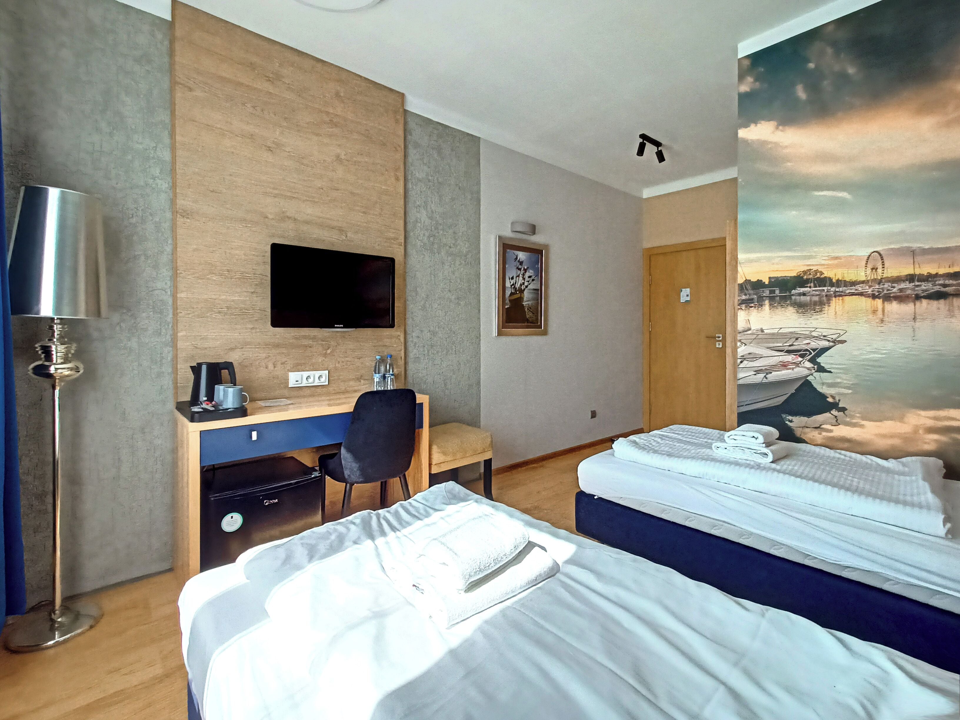 A hotel room with a desk, a flat-screen TV, a bed with two pillows, and a mural of a harbor scene.