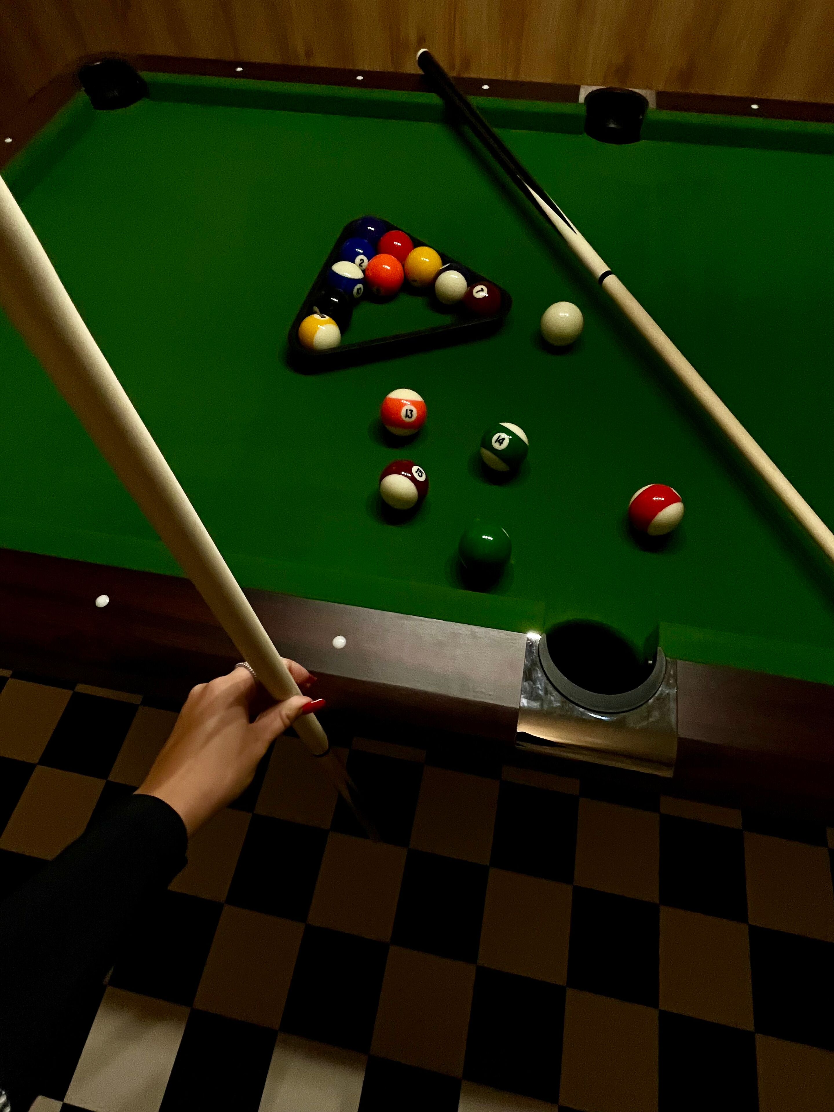 A pool table with balls and cues, a checkered floor, and a wooden wall.