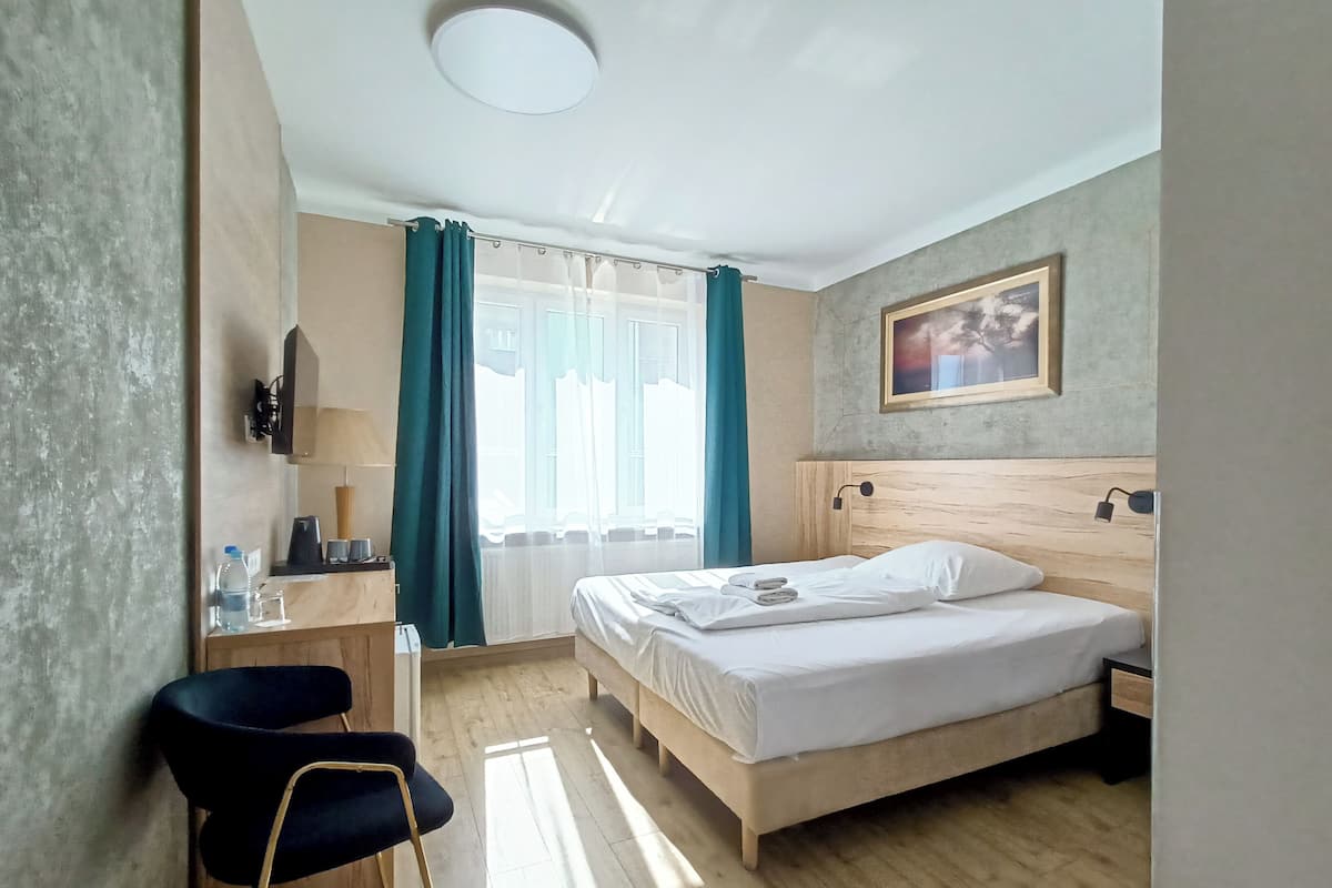 Une chambre d’hôtel avec un lit, un bureau, une chaise, une fenêtre avec des rideaux, un tableau au mur et un luminaire au plafond.