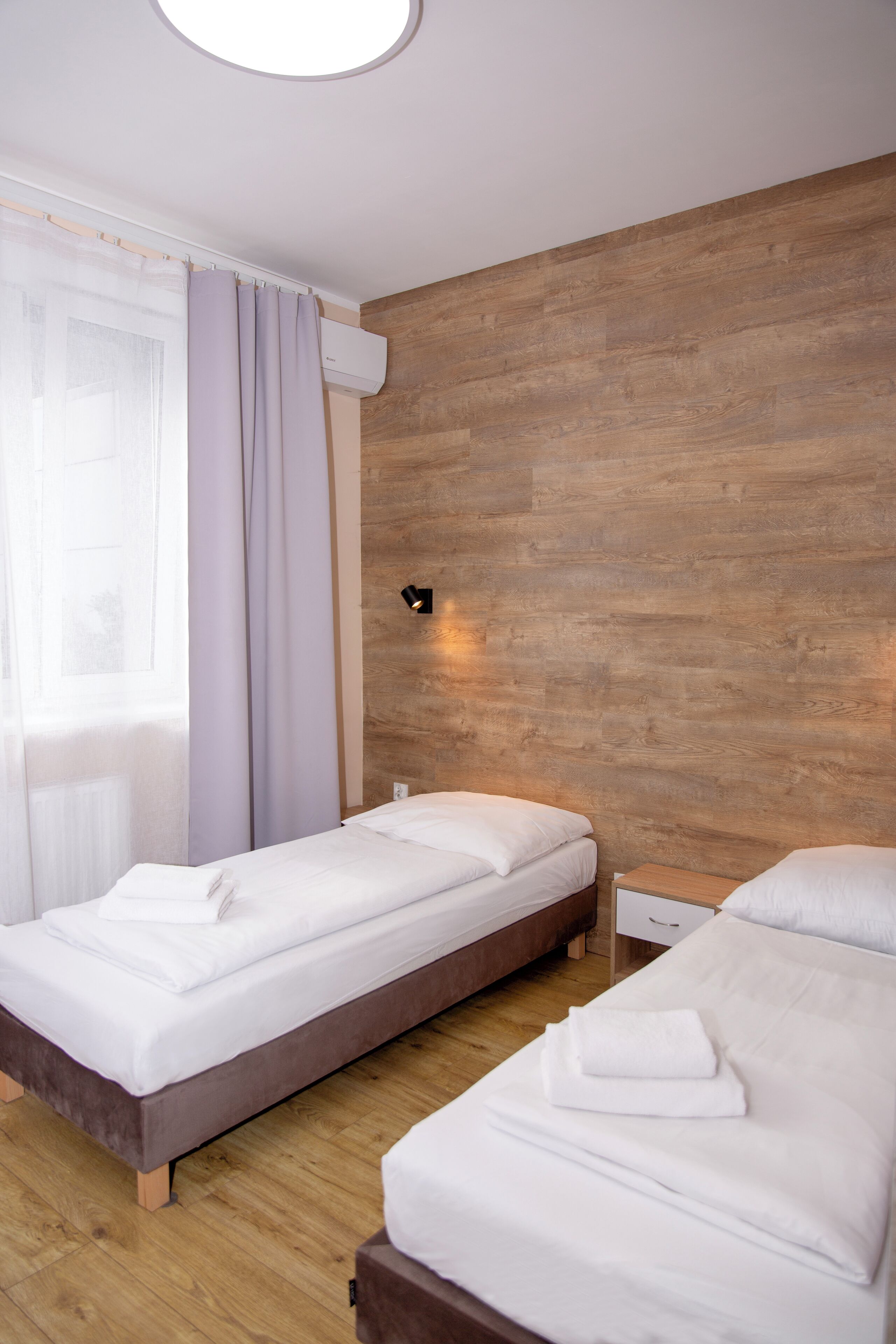 Ein Hotelzimmer mit zwei Betten, Holzboden, einem Fenster mit durchscheinenden Vorhängen und einer Wandleuchte.