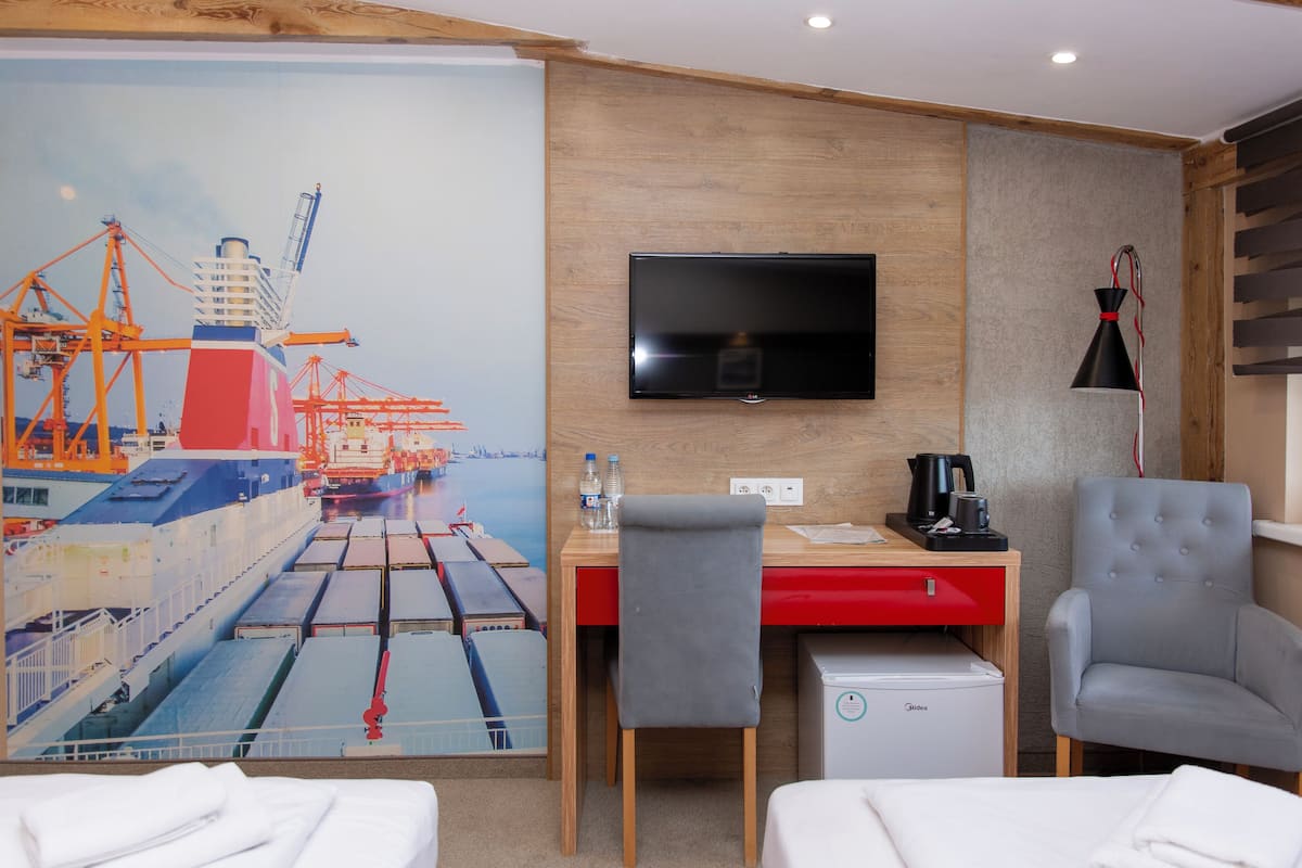 Une chambre d’hôtel avec un bureau en bois, une télévision, un petit réfrigérateur, un fauteuil gris et une fresque représentant un navire sur le mur.
