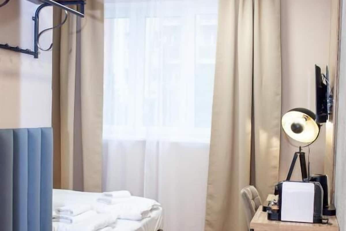 Une chambre d’hôtel de taille modeste, comprenant un lit, un bureau avec une lampe, une chaise et une fenêtre avec des rideaux.
