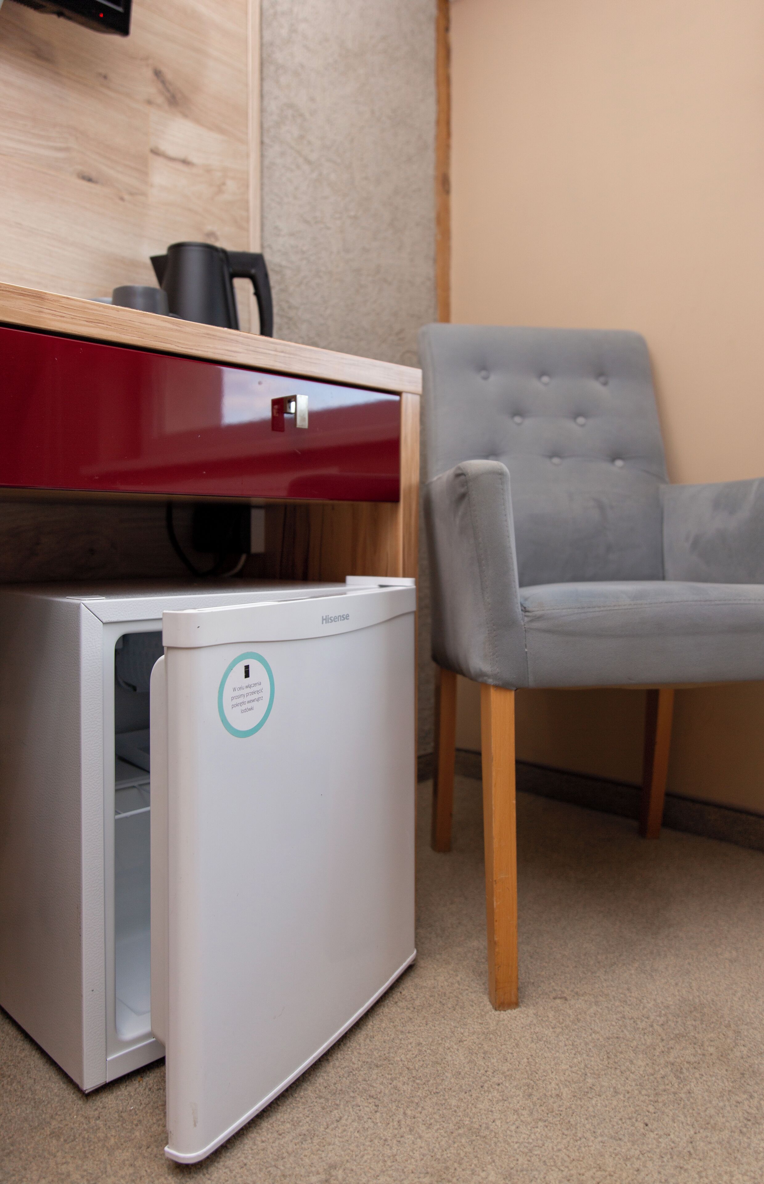 Un petit réfrigérateur blanc, fixé au mur, orné du logo Hisense, avec un plan de travail rouge au-dessus et une chaise rembourrée grise à sa droite.