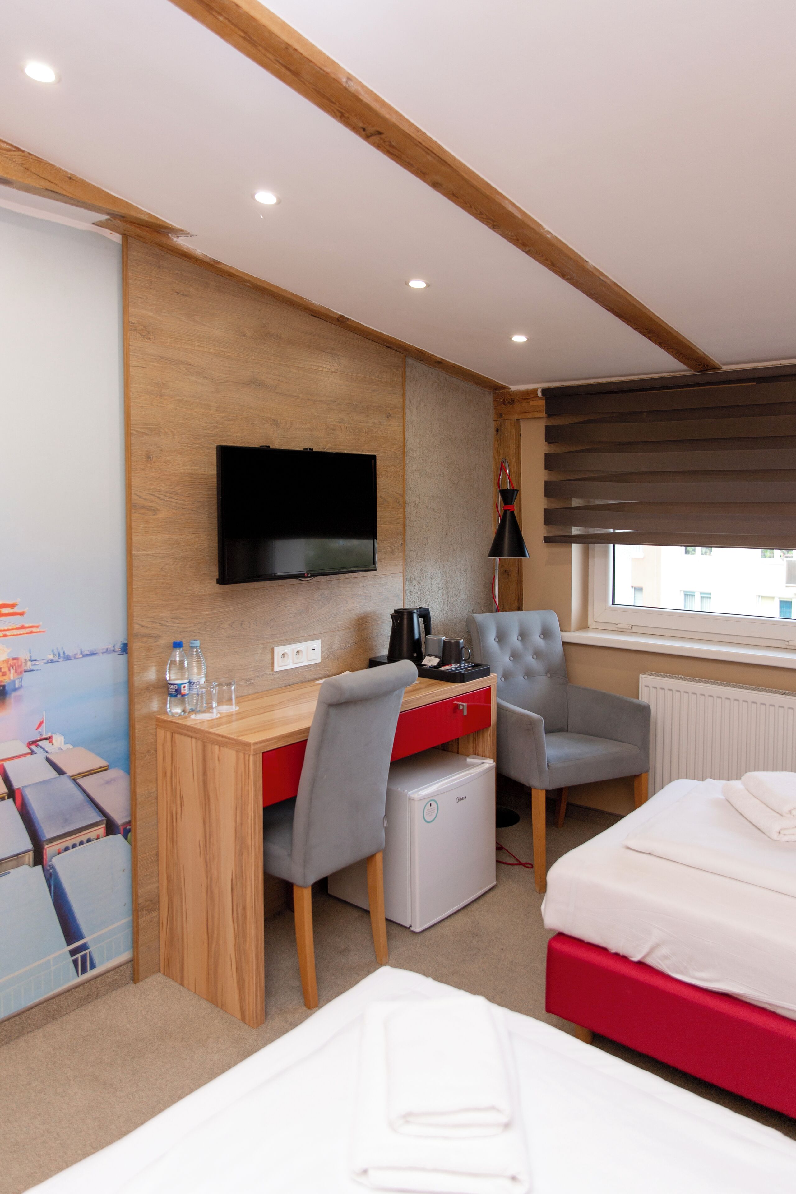 A modern hotel room with a wooden desk, a television, a bed, and a window with blinds.
