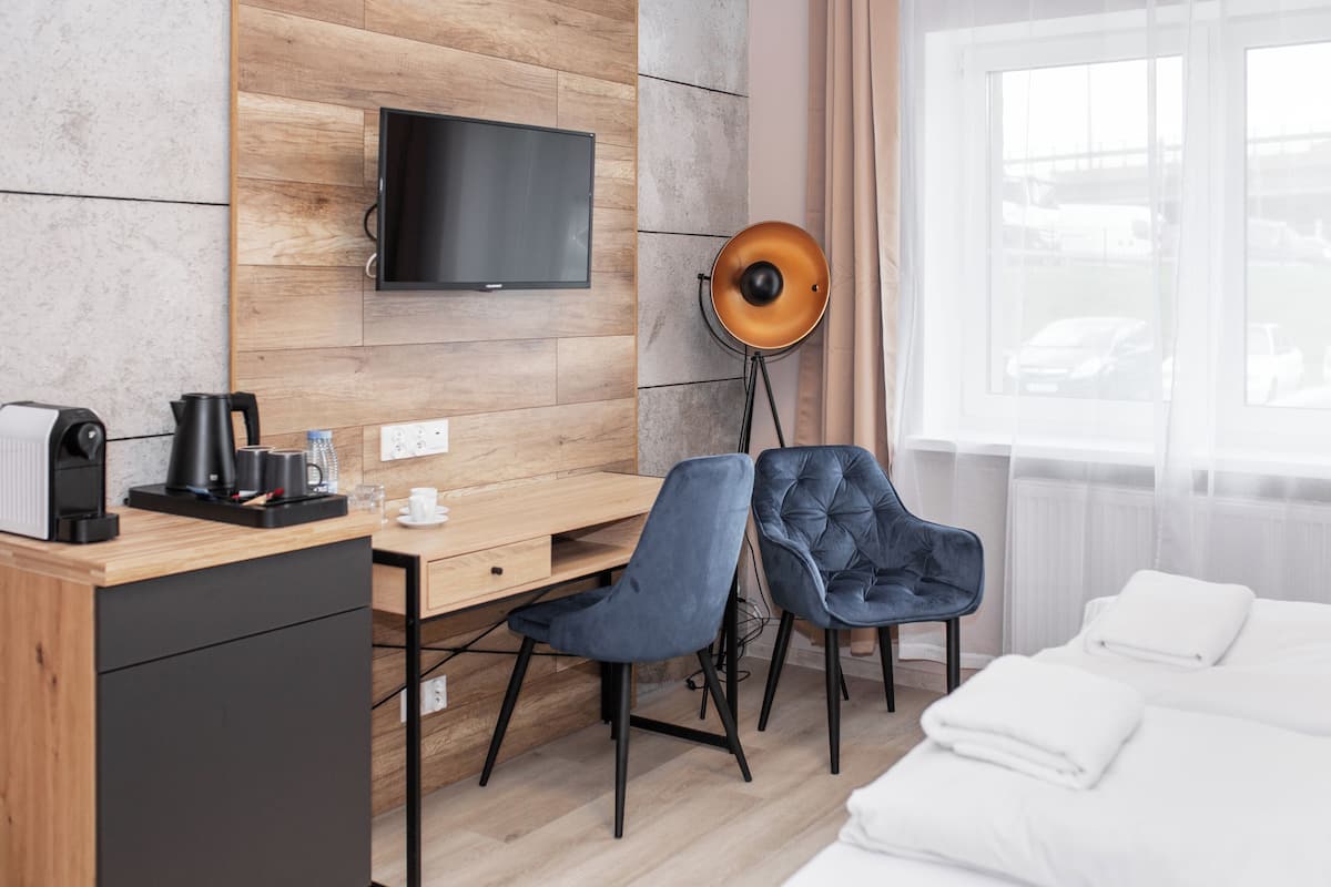 Une chambre d’hôtel avec un bureau en bois, un téléviseur fixé au mur, une lampe sur pied, deux chaises, un lit avec du linge de maison blanc et une fenêtre donnant sur des voitures garées.