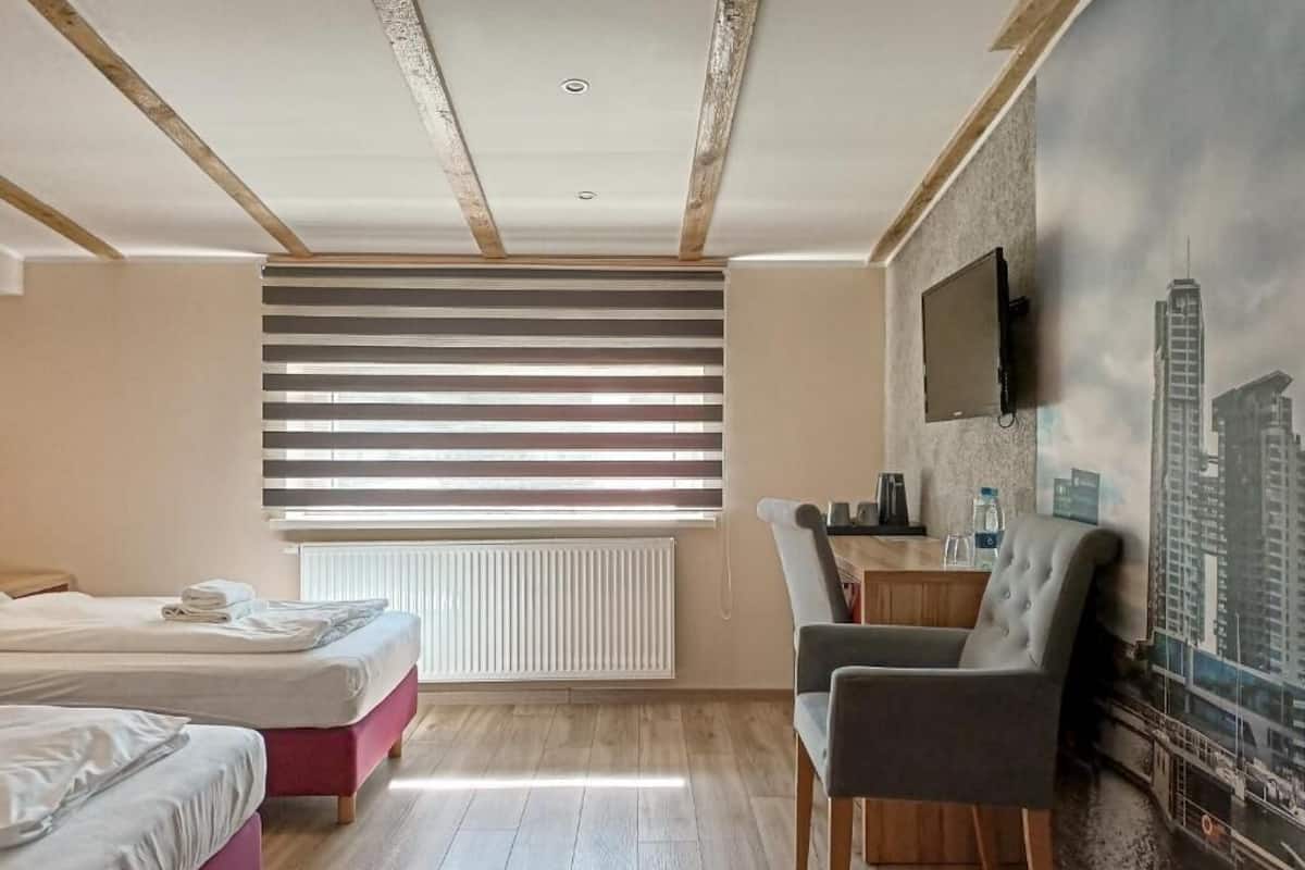 Une chambre d’hôtel avec deux lits, un bureau, une chaise, un radiateur et un tableau représentant un paysage urbain accroché au mur.