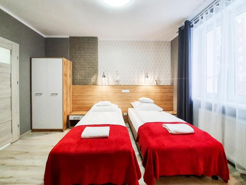 Quarto de hotel com duas camas de solteiro, um banco de madeira, um guarda-roupa e uma janela grande com cortinas.