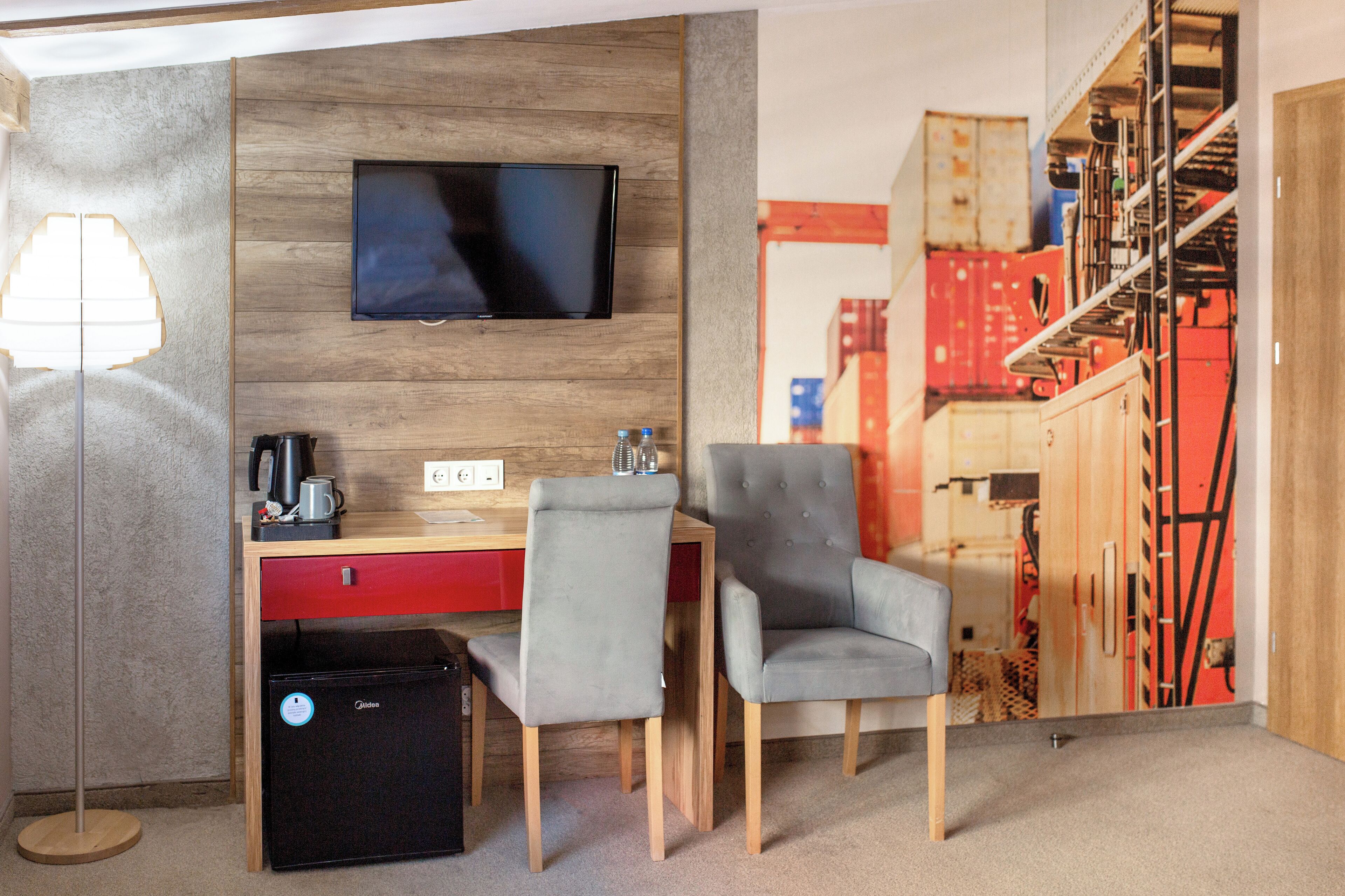 A hotel room with a wooden wall, a flat-screen TV, a desk with a red drawer, two grey chairs, a black suitcase, and a lamp.