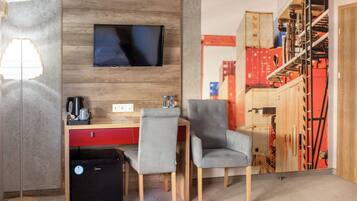 A hotel room with a wooden wall, a flat-screen TV, a desk with a red drawer, two grey chairs, a black suitcase, and a lamp.