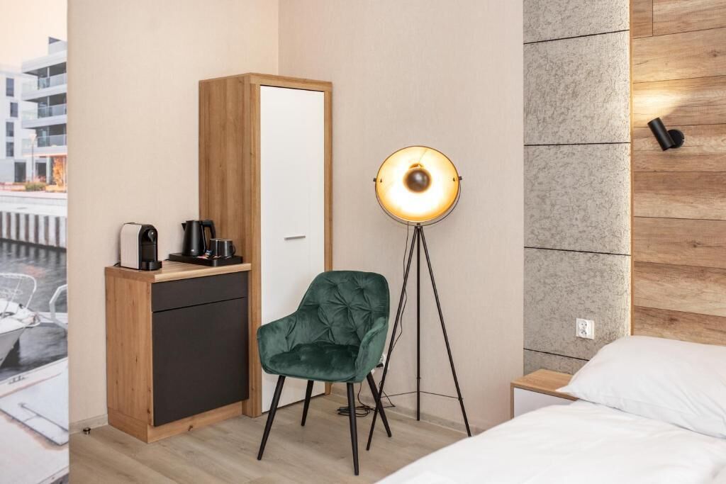 A hotel room with a wooden wardrobe, a green velvet chair, a floor lamp, and a bed with white bedding.