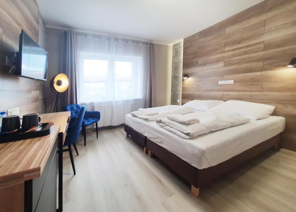 A modern hotel room with a wooden accent wall, a flat-screen TV mounted on the wall, a desk with a chair, a bed with white linens, and a window with curtains.