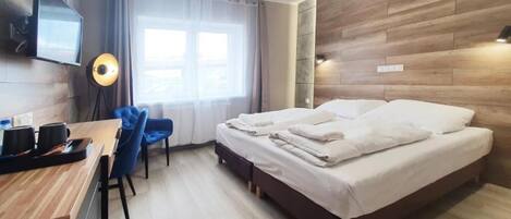A modern hotel room with a wooden accent wall, a flat-screen TV mounted on the wall, a desk with a chair, a bed with white linens, and a window with curtains.