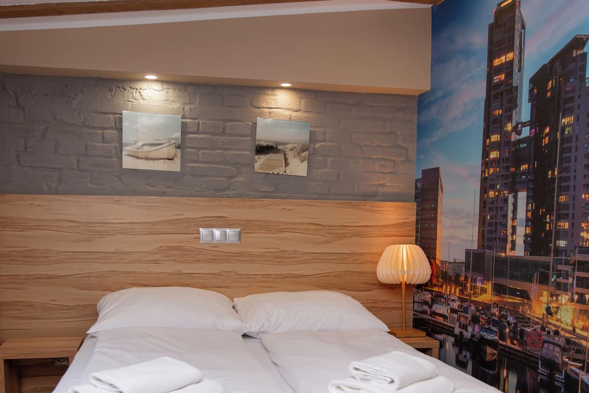 Un lit avec du linge de lit blanc et une base violette, une tête de lit en bois, un mur d’accent en pierre et une fresque représentant un paysage urbain.