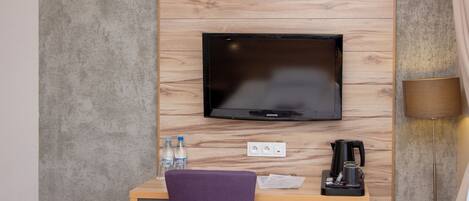 Habitación de hotel compacta con un escritorio de madera, un televisor montado en la pared, un refrigerador pequeño y una banca.